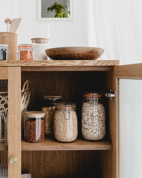 Photograph shows a selection of dried grains from a refill store displayed in glass jars in a wooden cupboard.